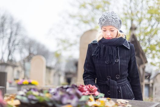 Solitary woman mourning with her hand on gravestone, remembering dead relatives in on Pere Lachaise cemetery in Paris, France.