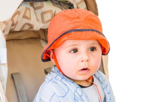 Baby with orange cap sitting in a stroller, looking amazed. Background isolated.