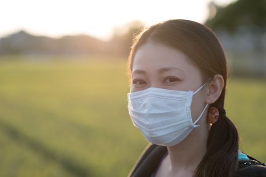 A young Japanese woman wearing a surgical mask outdoors in the countryside with a bright sunset behind her.