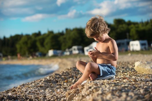 Cute little boy playing with his smart phone on the beach. Modern lifestyle, modern generation concept.