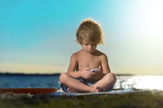 Portrait of a cute little boy playing with his smart phone on the beach. Modern lifestyle, modern generation concept.