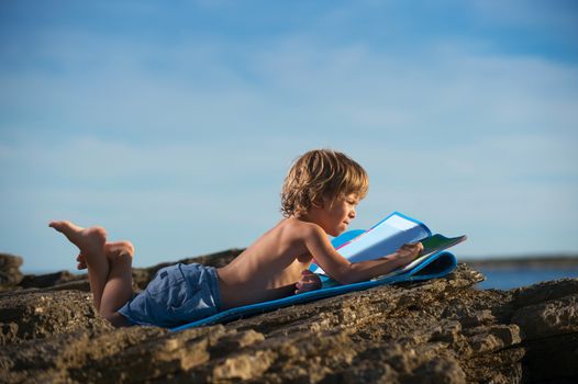 Cute little boy lying on his stomach looking at the picture book on the beach. Somen negative space around.