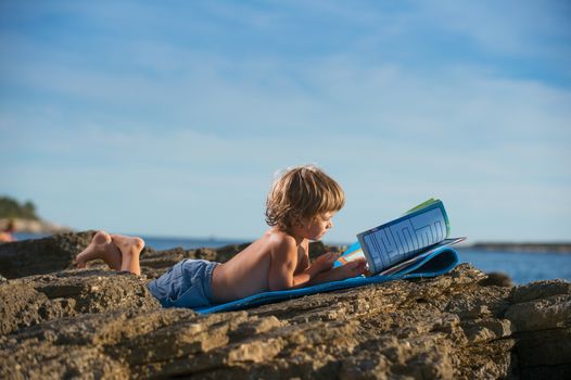 Cute little boy lying on his stomach reading book on the beach. Some negative space around.