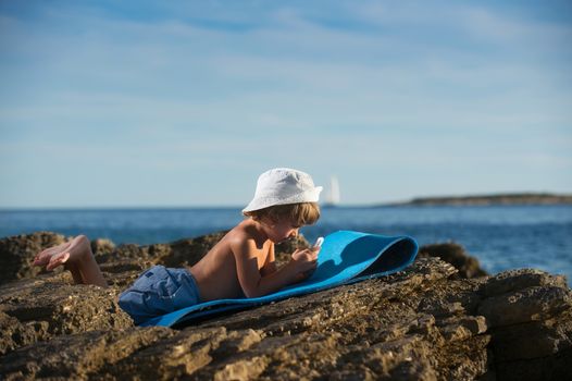 Cute little boy with a cap lying on the beach playing with his smart phone. Modern lifestyle, modern generation concept.
