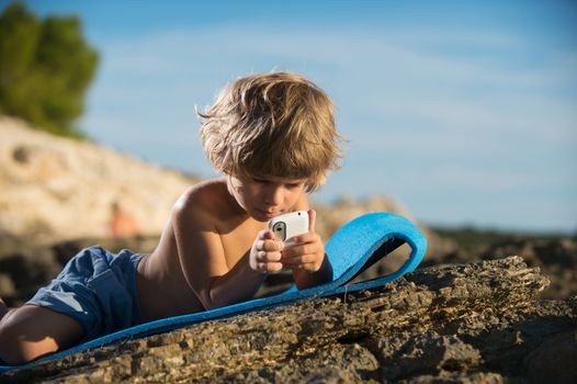 Cute little boy lying on the beach playing with his smart phone. Modern lifestyle, modern generation concept.