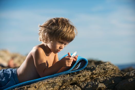 Cute little boy lying on the beach playing with his smart phone. Modern lifestyle, modern generation concept.