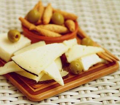 Arrangement of Goat Cheeses with Bread Sticks and Green Olives on Wooden Plate closeup on Wicker background. Focus on Foreground
