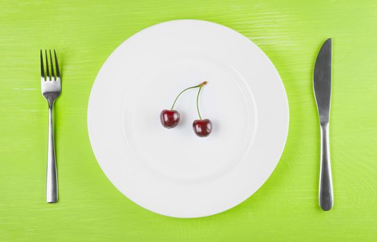 The concept of a healthy lifestyle, diet, weight loss, anti-obesity, healthy diet, raw food diet, vegetarianism, veganism. Two ripe cherries on a white plate, knife and fork, top view, closeup