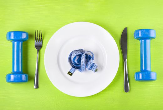 The concept of a healthy lifestyle, diet, sports, weight loss, anti-obesity, exercise, healthy diet.  Centimeter on a plate, knife, dumbbells and fork, top view, closeup