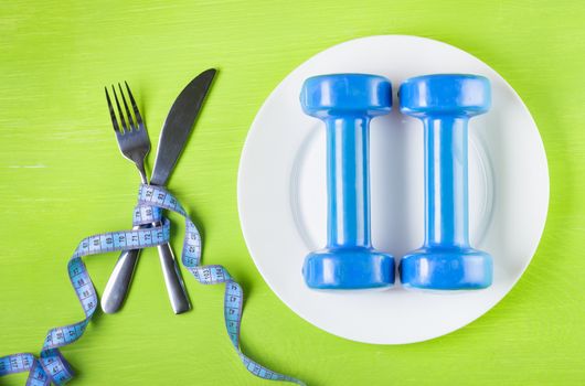 The concept of a healthy lifestyle, diet, sports, weight loss, anti-obesity, exercise, healthy diet.   Dumbbells on a plate, crossed fork and knife wrapped centimeter on a green table top view, 