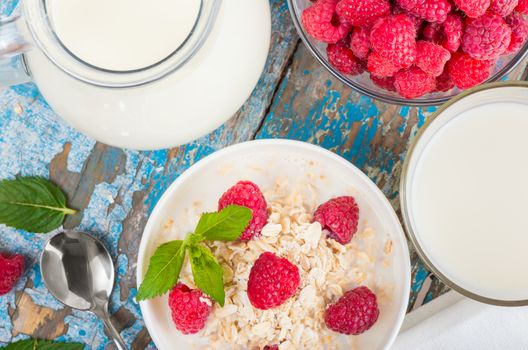 Oat flakes with milk and raspberries for breakfast Jug and glass with milk, spoon, napkin, fresh mint on an old wooden blue background, top view. The concept of a healthy diet, weight loss