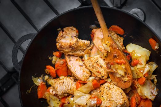 Roasted turkey fillet with vegetables in a frying pan on a plate, a wooden spoon mixes grilled meats, top view, close up  