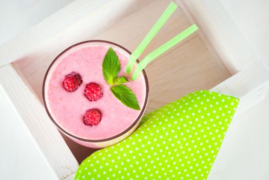 Raspberry dairy smoothie with fresh berries and sprig of mint with two green cocktail stick in a decorative box on a white wooden table