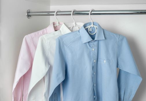 white, blue and pink clean ironed men's shirts hanging on hangers in the white wardrobe