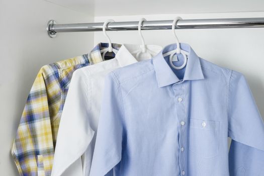 white, blue and checkered clean ironed men's shirts hanging on hangers in the white wardrobe 