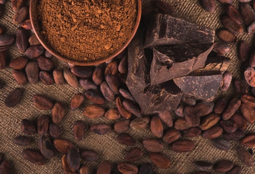 Raw cocoa beans, clay bowl with cocoa powder, black chocolate on brown sacking, top view