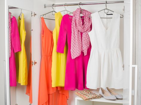 bright colorful female dresses hanging on coat hanger, pink scarf, shoes and handbag in white wardrobe