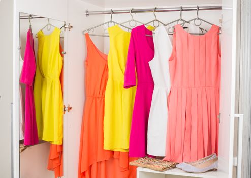 bright colorful female dresses hanging on coat hanger,  shoes and handbag in white wardrobe