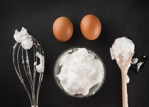Whipped egg whites for cream in a glass bowl, whisk and wooden spoon on a dark background, top view