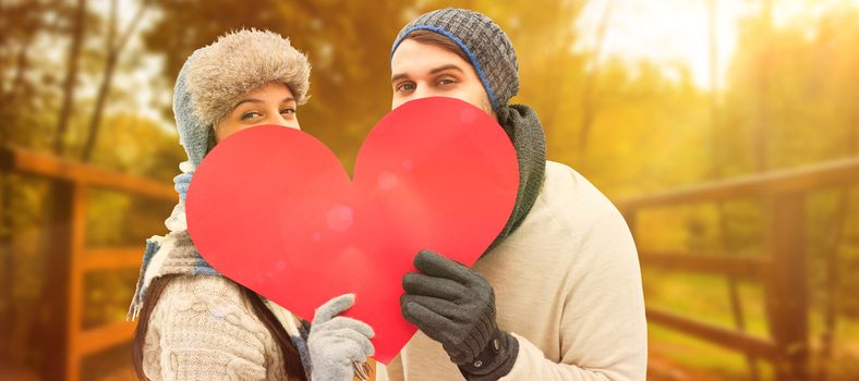 Attractive young couple in warm clothes holding red heart against autumn scene