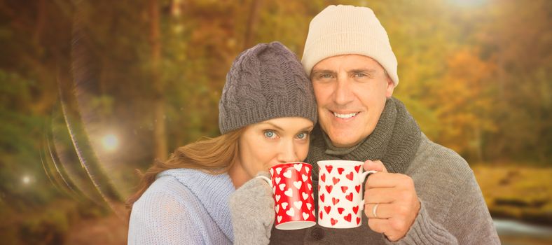 Happy couple in warm clothing holding mugs against autumn scene