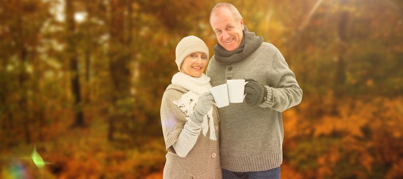 Happy mature couple in winter clothes holding mugs against autumn scene