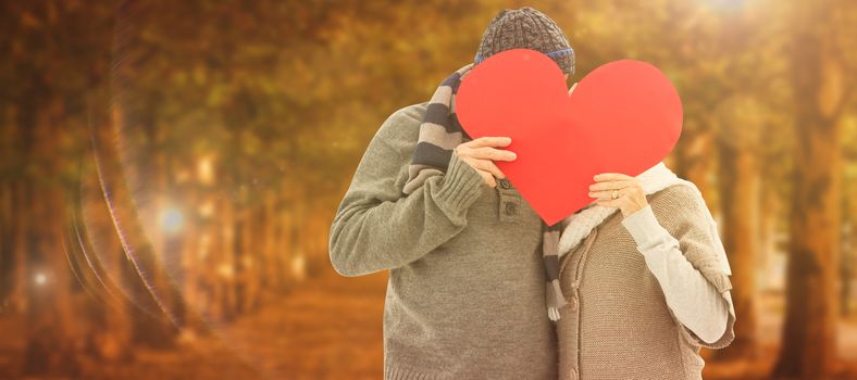 Happy mature couple in winter clothes holding red heart against autumn scene