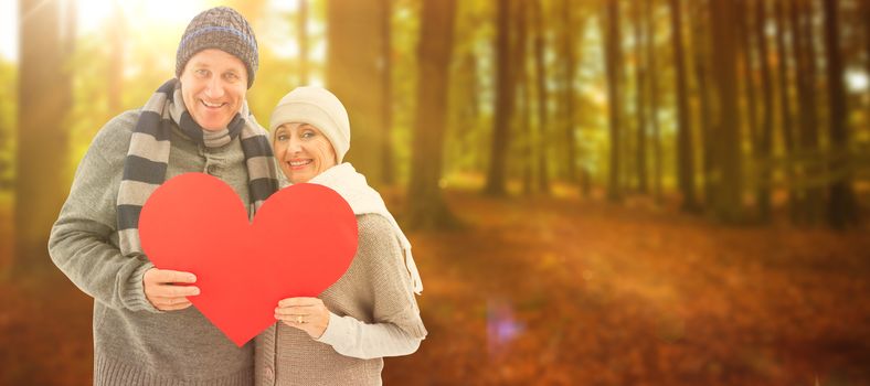 Happy mature couple in winter clothes holding red heart against autumn scene