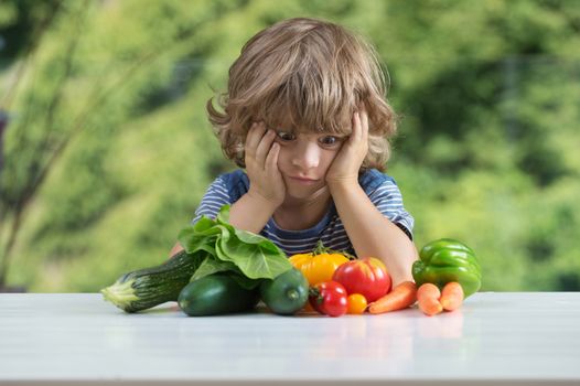 Cute little boy sitting at the table, unhappy with his vegetable meal, bad eating habits, nutrition and healthy eating concept