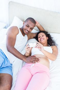 Pregnant wife holding smartphone with husband lying on bed at home