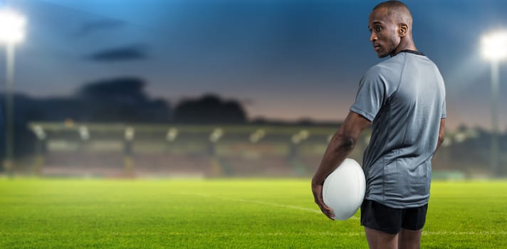 Rear view of confident athlete standing with rugby ball against pitch