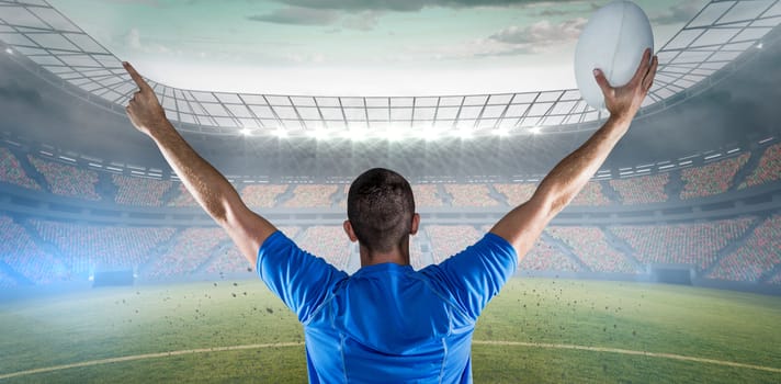 Rear view of rugby player holding ball with arms raised against rugby stadium