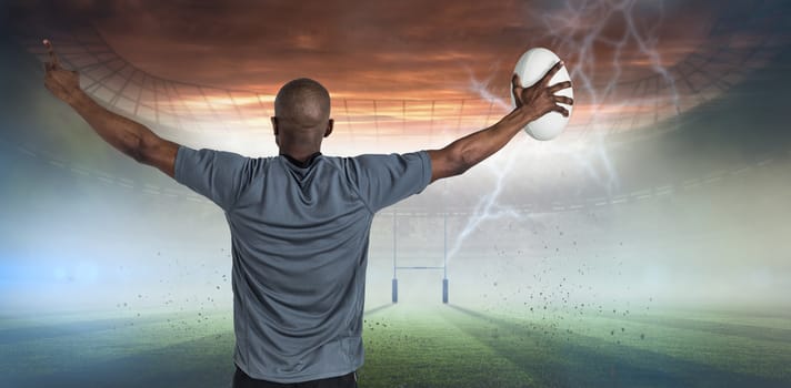 Rear view of sportsman with arms raised holding rugby ball against rugby pitch
