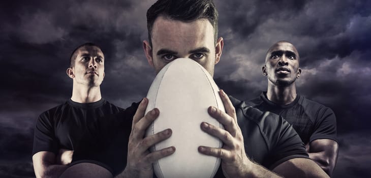 Tough rugby player holding ball against blue sky with white clouds