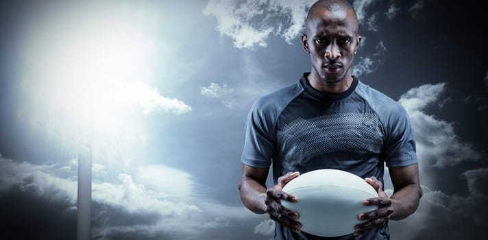 Portrait of serious sportsman holding rugby ball against spotlight in sky