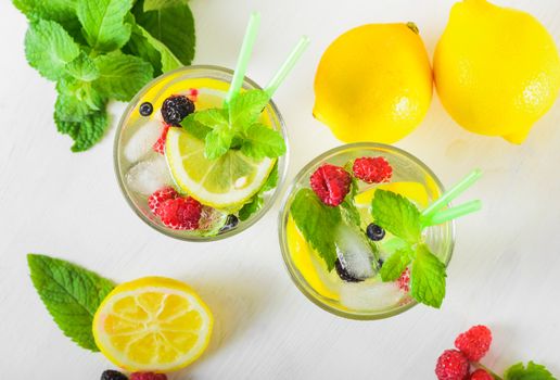 Delicious refreshing lemonade with fresh mint, Lemon, honey and ice decorated with raspberries, blueberries and blackberries on a white wooden table, close up. Healthy summer drink. Top view