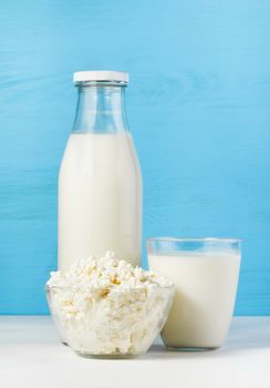 tasty healthy dairy products on a white table on a blue background:  cottage cheese in bowl,  milk in a glass bottle and in a glass