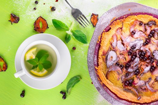 Good morning, homemade Crumble pie with black currants, plums and powdered sugar for breakfast, mint tea with lemon, fork, whisk, dried plum, mint leaves on wooden green table, top view, composition