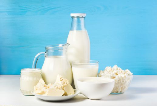 tasty healthy dairy products on a white table on a blue background: sour cream in a white bowl, cottage cheese in bowl, cream in a a bank,  butter on a saucer and milk in a jar