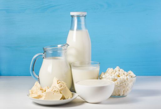 tasty healthy dairy products on a white table on a blue background: sour cream in a white bowl, cottage cheese in bowl, butter on a saucer and milk in a jar, glass bottle and in a glass
