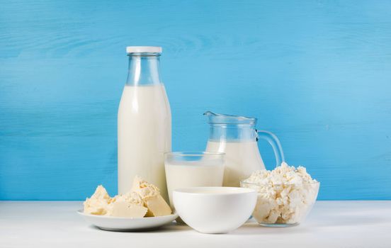 tasty healthy dairy products on a white table on a blue background: sour cream in a white bowl, cottage cheese in bowl,  butter on a saucer, milk in a jar, glass bottle and in a glass