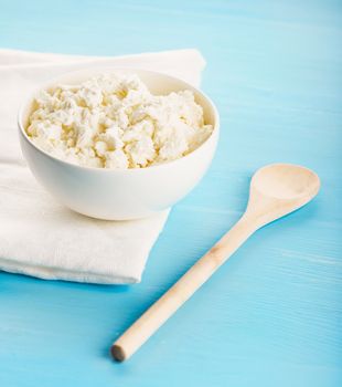 Tasty healthy village cottage cheese in a white ceramic bowl, a white kitchen cloth and wooden spoon on a blue wooden table