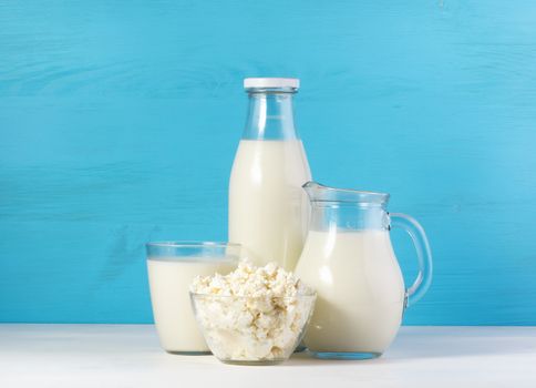 tasty healthy dairy products on a white table on a blue background: cottage cheese in bowl, milk in a jar, glass bottle and in a glass