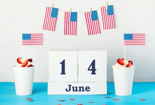 Greeting card on the day of the American flag, two desserts with cream, strawberries and American flags, perpetual calendar with the words June 14 , American flags hanging on a rope with clothespins 