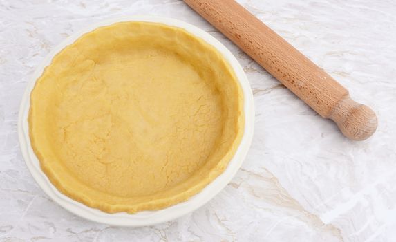 Ceramic pie dish lined with fresh shortcrust pastry on a kitchen counter with a wooden rolling pin