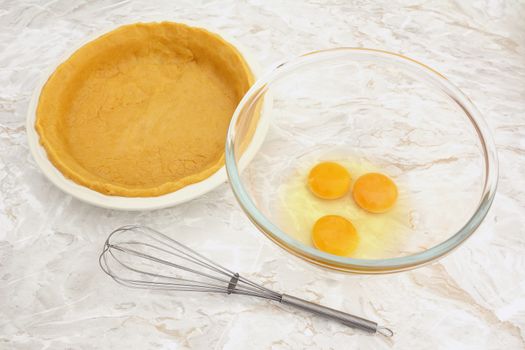 Bowl of raw eggs with a wire whisk and a pie dish lined with shortcrust pastry
