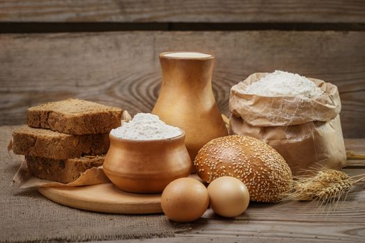 Ingredients for baking bread and pastry, milk, wheat flour, eggs, bun with sesame, rustic bread, cut into pieces, ears of wheat on the old wooden background