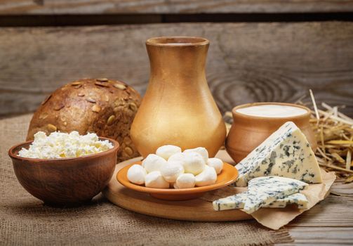 Rustic natural dairy products, whole milk, cottage cheese, cut slices of blue cheese with mold, mozzarella cheese, rustic bread, burlap, straw on the old wooden background