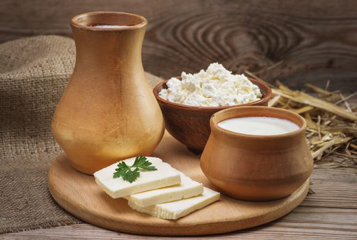 Rustic natural dairy products, whole milk in a clay jug and a cup, cottage cheese in a bowl, cut slices of cheese, burlap, straw on the old wooden background