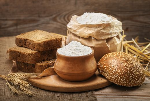 Wheat flour, sliced bread, bun with sesame seeds, wheat ears and straw on an old wooden background